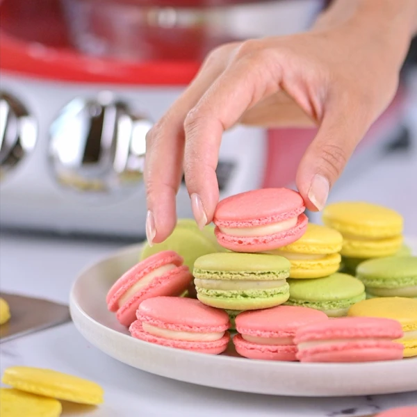 Hands holding pink and green macarons in a kitchen setting.