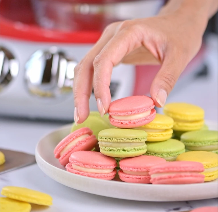 Händer som håller rosa och gröna macarons i en köksmiljö.